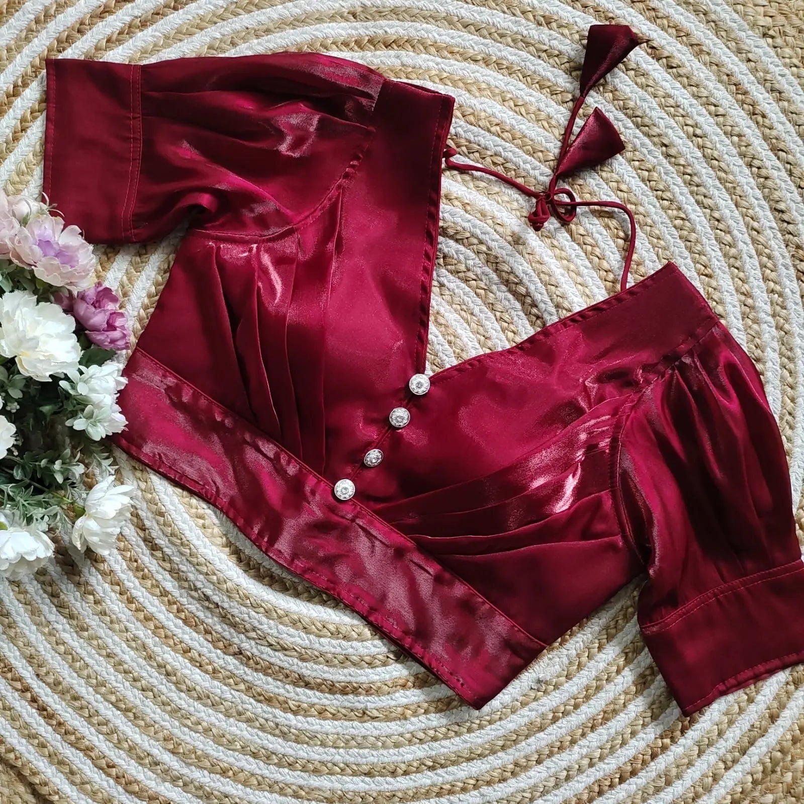 Maroon satin blouse with pleated detailing and four decorative silver buttons on the front, featuring elbow-length sleeves and tie-up tassels, displayed on a circular woven mat with white and pink flowers on the side.