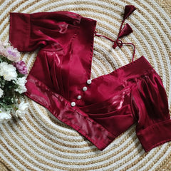 Maroon satin blouse with pleated detailing and four decorative silver buttons on the front, featuring elbow-length sleeves and tie-up tassels, displayed on a circular woven mat with white and pink flowers on the side.