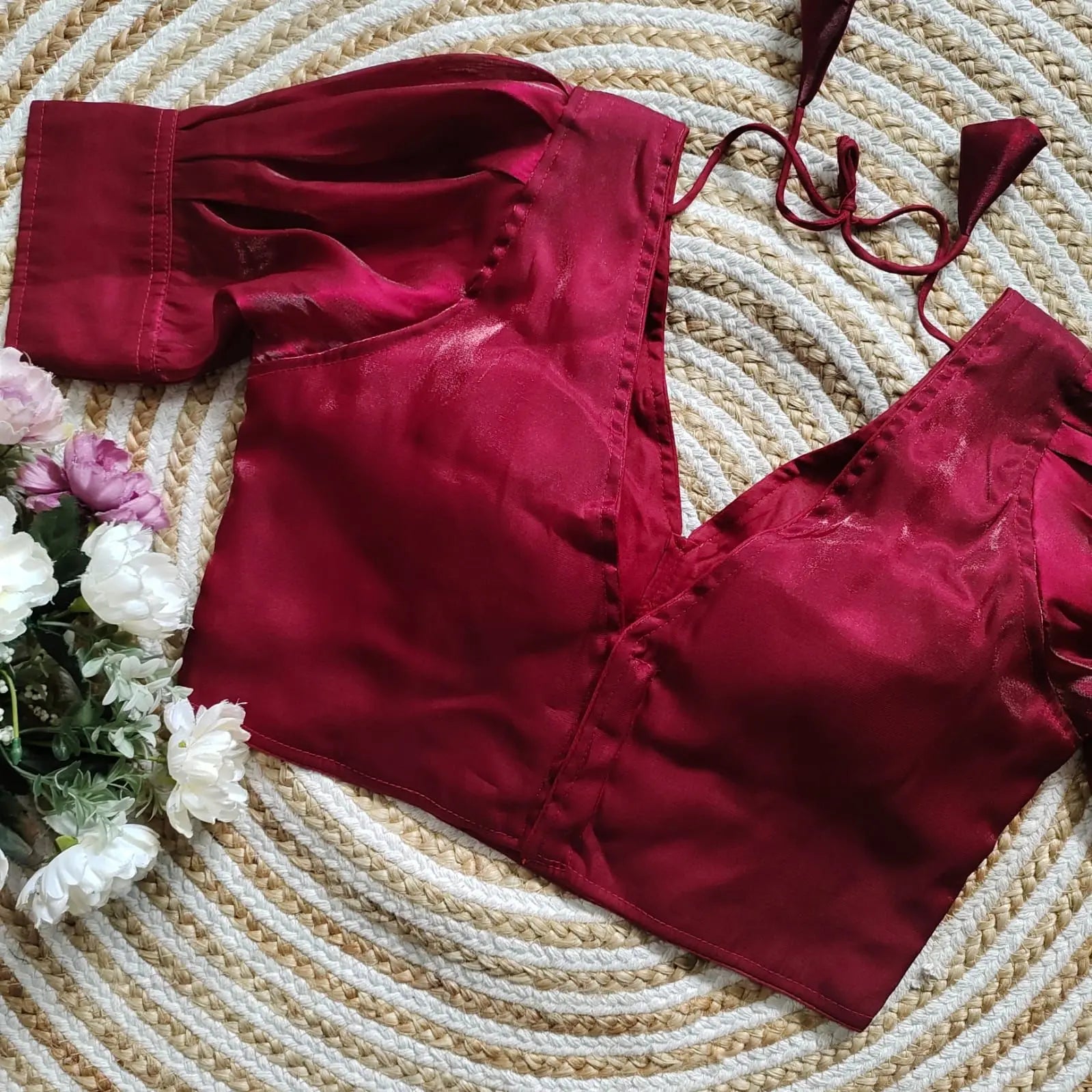 Maroon satin blouse with pleated detailing and four decorative silver buttons on the front, featuring elbow-length sleeves and tie-up tassels, displayed on a circular woven mat with white and pink flowers on the side.