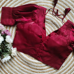 Maroon satin blouse with pleated detailing and four decorative silver buttons on the front, featuring elbow-length sleeves and tie-up tassels, displayed on a circular woven mat with white and pink flowers on the side.