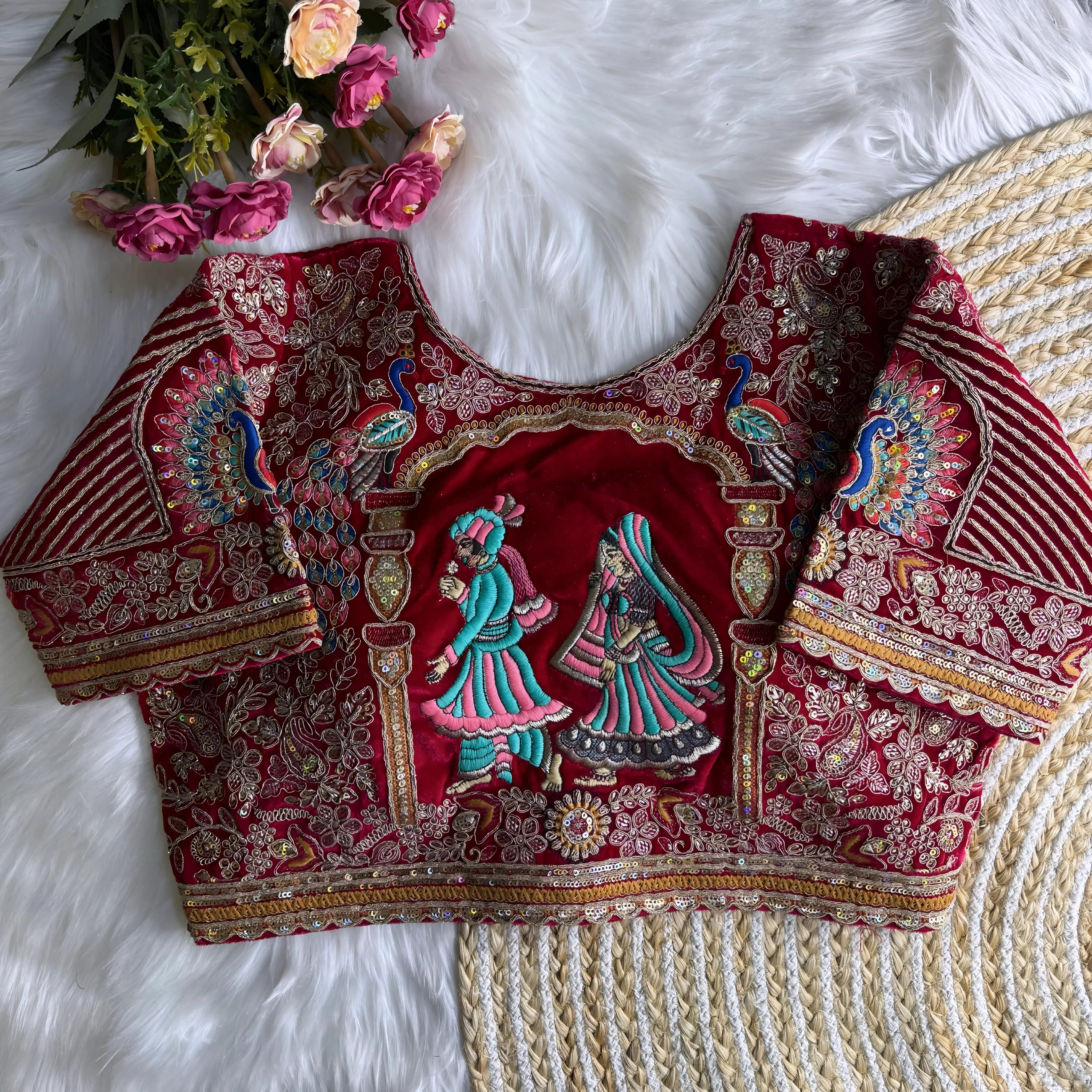 Maroon silk blouse featuring intricate gold zardosi embroidery with floral and paisley motifs, sequin polka dot accents, striped geometric panels, and a colorful peacock feather design on the sleeve, displayed flat on a white fur surface with a braided mat and pink artificial flowers in the corner.