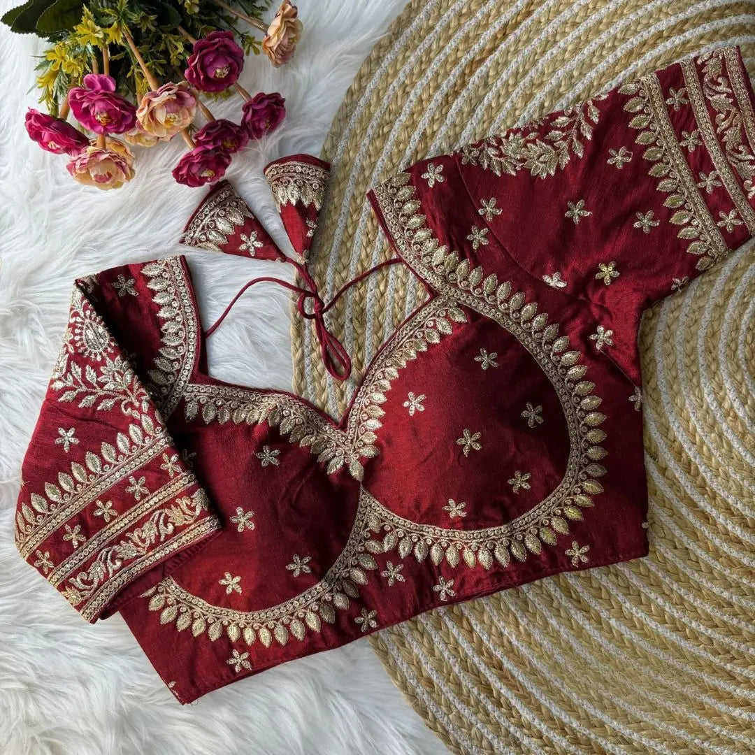 Maroon blouse with intricate gold zari embroidery, featuring floral and leaf motifs, decorative borders on sleeves and neckline, and tie-up tassels, displayed on a textured mat with pink flowers in the background.