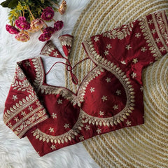 Maroon blouse with intricate gold zari embroidery, featuring floral and leaf motifs, decorative borders on sleeves and neckline, and tie-up tassels, displayed on a textured mat with pink flowers in the background.