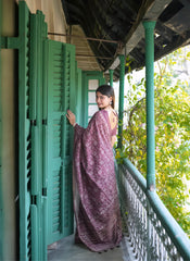 A woman models an elegant Wine Red/Deep Maroon Tussar Silk Saree. The saree features dense all-over floral embroidery/weaving in a light metallic color, framed by a contrasting light metallic border. She wears a matching wine short-sleeve blouse and subtle silver bangles, posing on a balcony framed by vibrant teal shutters.