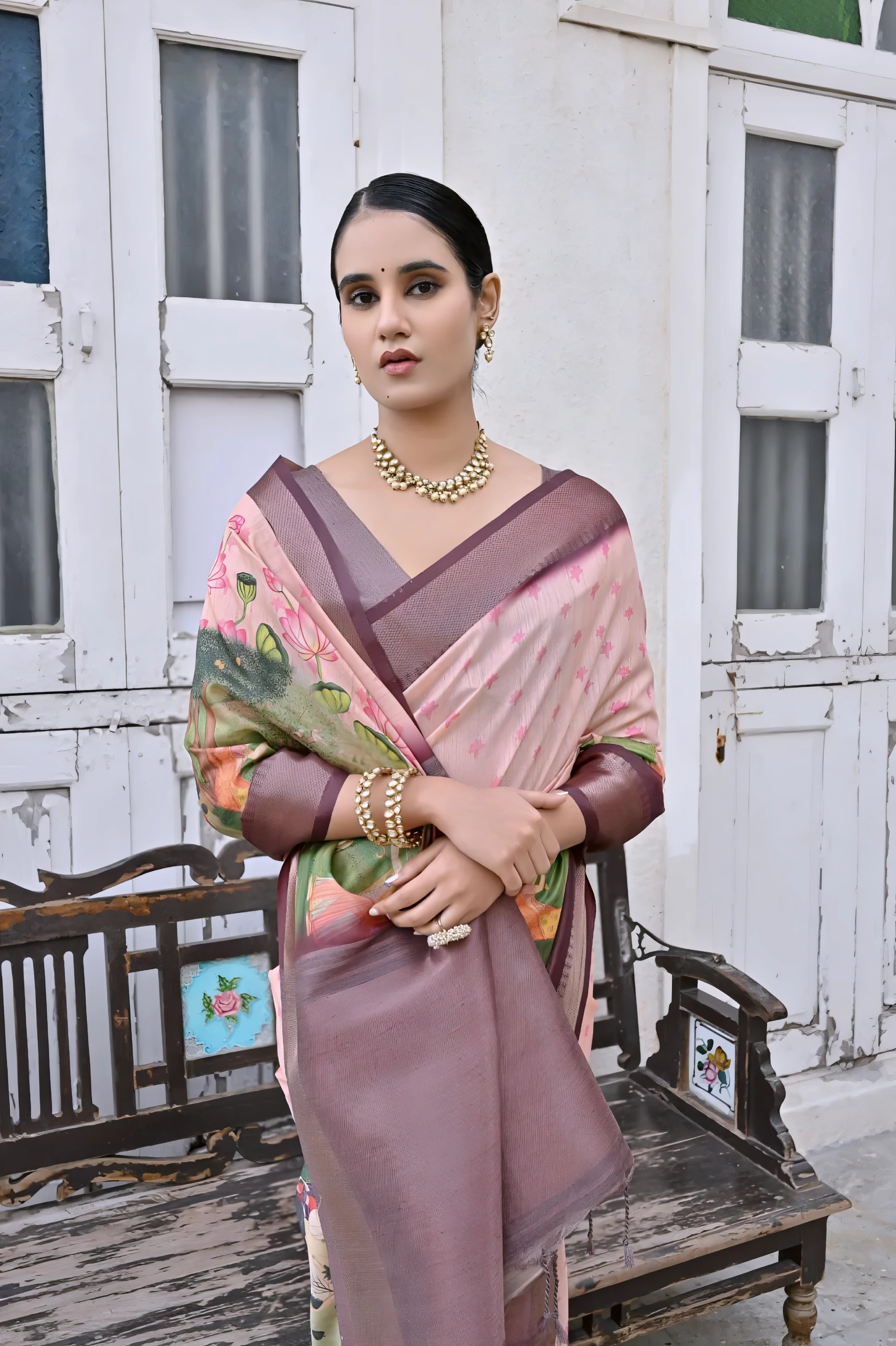 A woman models an elegant Light Peach Soft Silk Saree featuring a large-scale floral and figurative Pichwai-style print in soft colors. The saree has a wide Deep Brown/Maroon border and pallu. She wears a matching dark blouse and a gold Kundan choker necklace, posing outdoors next to distressed antique white doors.