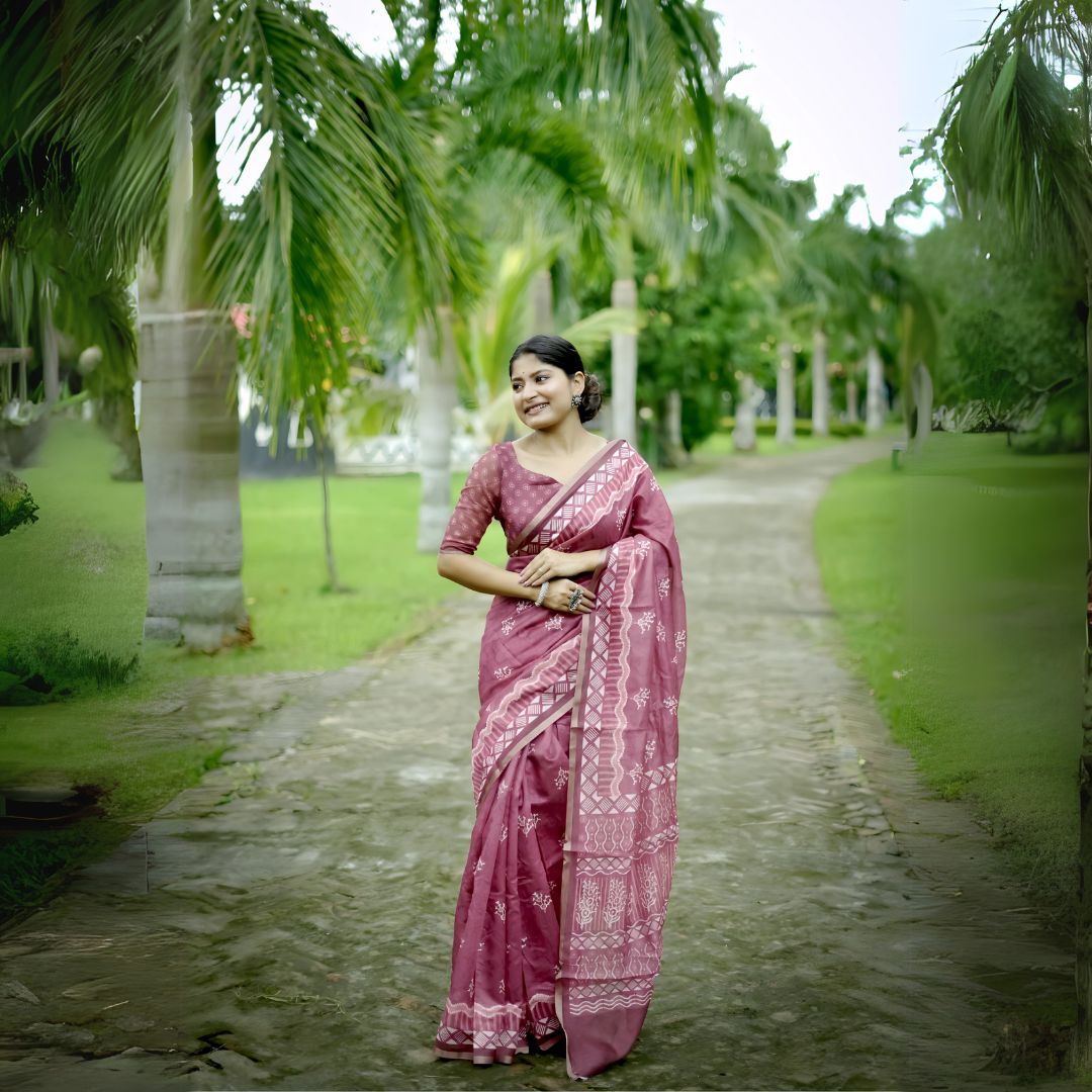 A woman models a Dusty Rose Pink Soft Cotton Saree featuring an all-over digital block print of traditional motifs. She wears a sheer-sleeved pink blouse and silver jhumka earrings, posing on an outdoor path lined with palm trees.