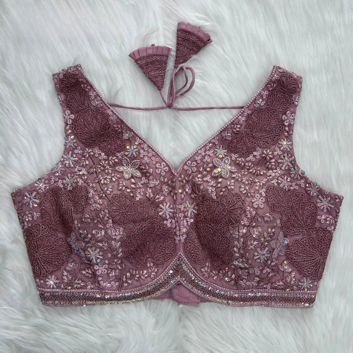Mauve blouse featuring intricate floral sequin and thread embroidery with large lace appliqué motifs, sparkling bead accents, and a scalloped border, deep V-neckline with structured cups, and pleated fan-designed tasseled tie-back, displayed flat on a soft white fur background.