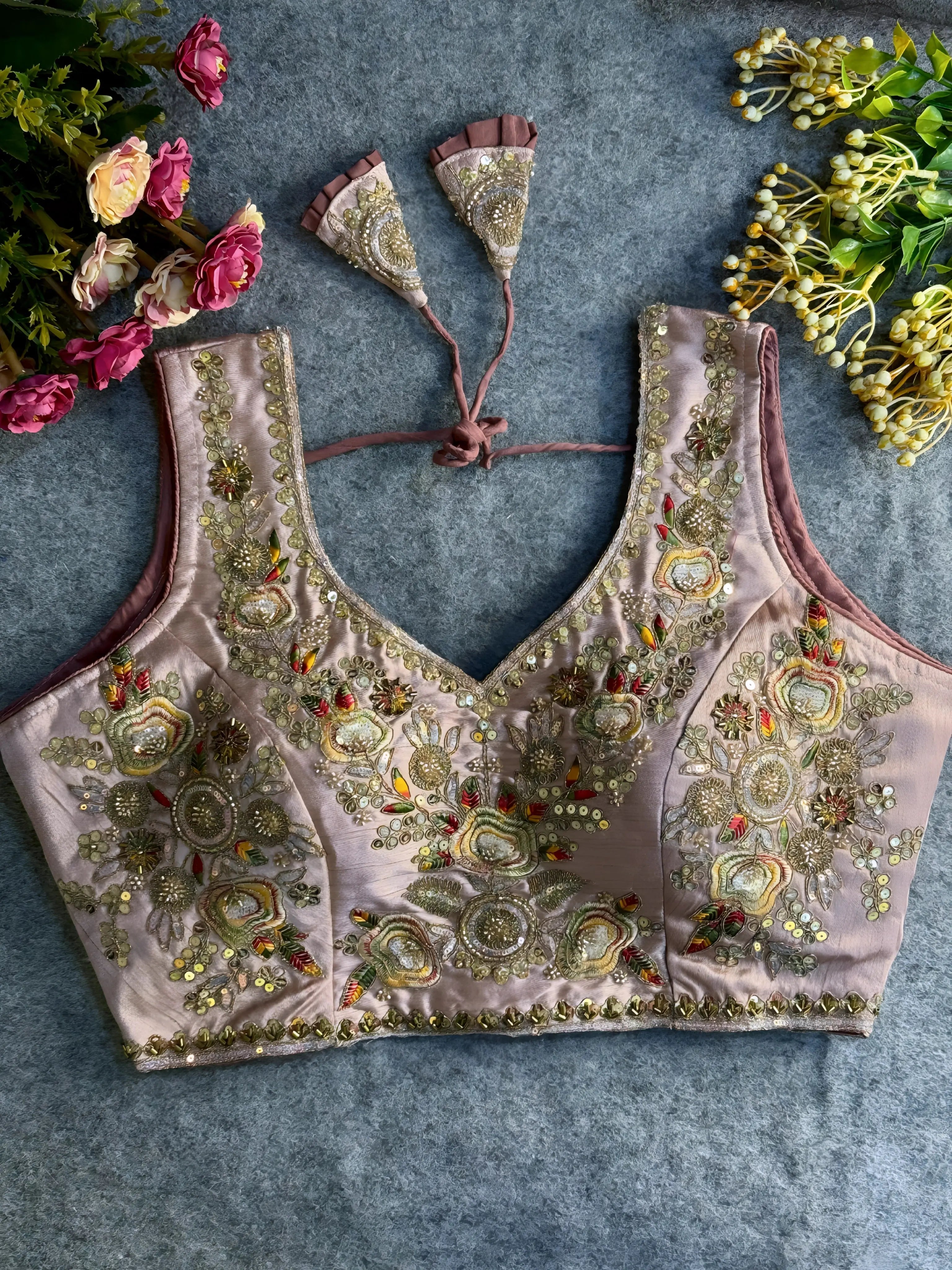 Traditional mauve silk blouse featuring intricate gold zardozi embroidery and beadwork with multicolor floral and leaf motifs on the front and neckline, finished with decorative tassel ties, displayed flat on a textured grey background with artificial pink and yellow flowers in the corners.