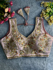 Traditional mauve silk blouse featuring intricate gold zardozi embroidery and beadwork with multicolor floral and leaf motifs on the front and neckline, finished with decorative tassel ties, displayed flat on a textured grey background with artificial pink and yellow flowers in the corners.