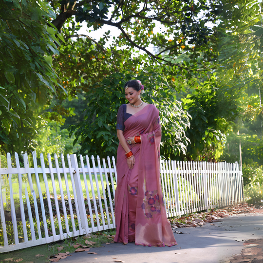 A woman models an elegant Dusty Rose Pink Soft Muga Cotton Saree. The pallu features large, colorful Resham floral weaving in purple and dark pink. She wears a slate grey blouse and is accessorized with a pearl choker necklace and vibrant orange bangles, posing outdoors by a white fence.