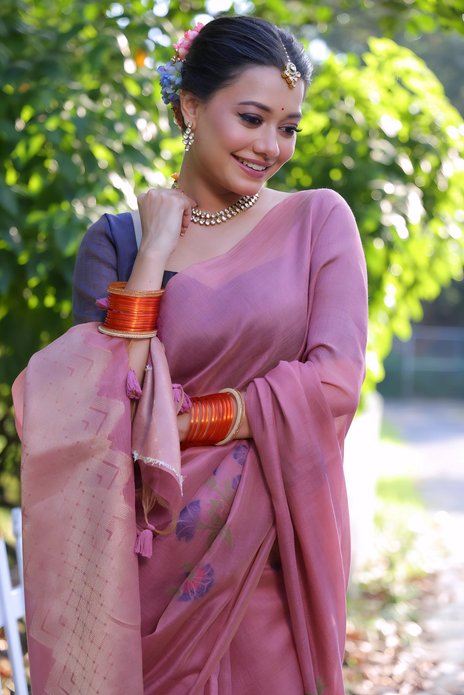 A woman models an elegant Dusty Rose Pink Soft Muga Cotton Saree. The pallu features large, colorful Resham floral weaving in purple and dark pink. She wears a slate grey blouse and is accessorized with a pearl choker necklace and vibrant orange bangles, posing outdoors by a white fence.