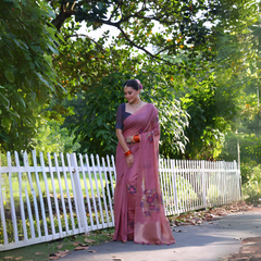A woman models an elegant Dusty Rose Pink Soft Muga Cotton Saree. The pallu features large, colorful Resham floral weaving in purple and dark pink. She wears a slate grey blouse and is accessorized with a pearl choker necklace and vibrant orange bangles, posing outdoors by a white fence.