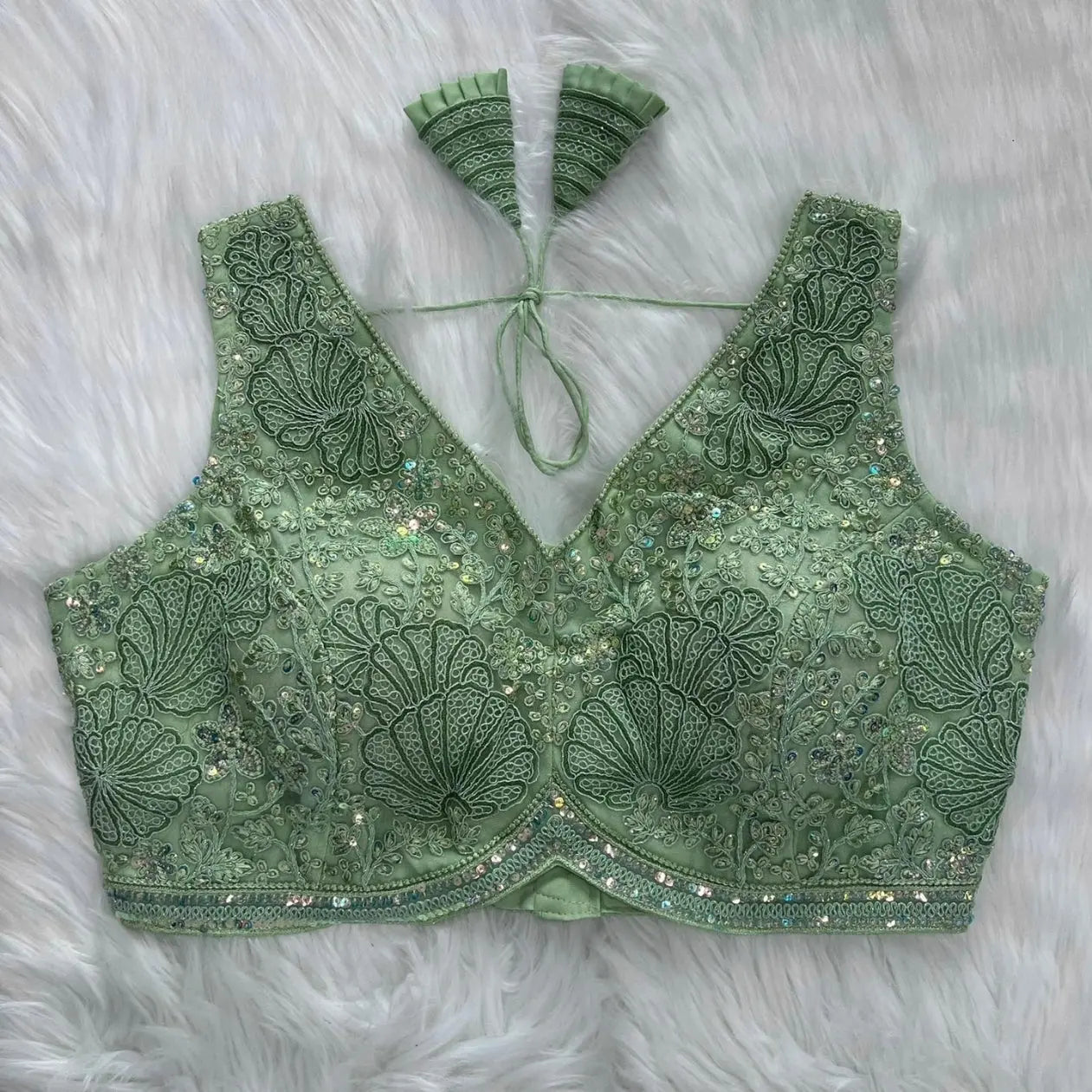 Mint green blouse featuring intricate shell-shaped embroidery, sequin and bead embellishments, floral vine motifs, and a decorative border, with a deep V-neck, structured cups, and pleated fan-style tasseled tie-back, displayed flat on a white fur background.