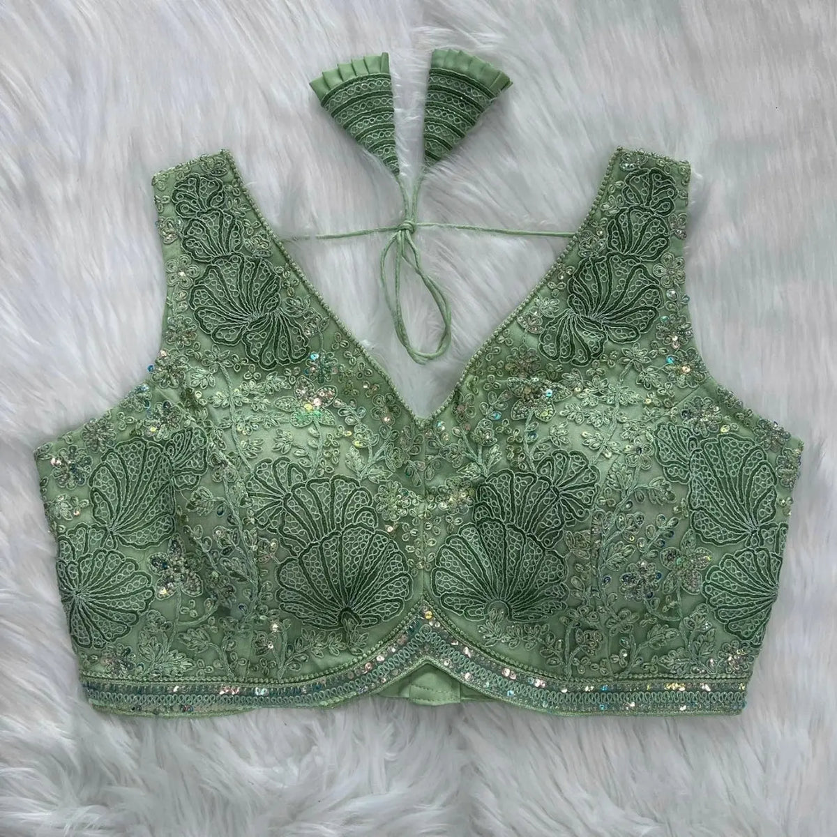 Mint green blouse featuring intricate shell-shaped embroidery, sequin and bead embellishments, floral vine motifs, and a decorative border, with a deep V-neck, structured cups, and pleated fan-style tasseled tie-back, displayed flat on a white fur background.