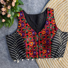 Sleeveless black ethnic blouse highlighted by vibrant multicolored mirror-work embroidery on the front, metallic chevron side panels, and cowrie-shell tassels along the hem, laid on a grey backdrop beside a woven mat and floral greenery