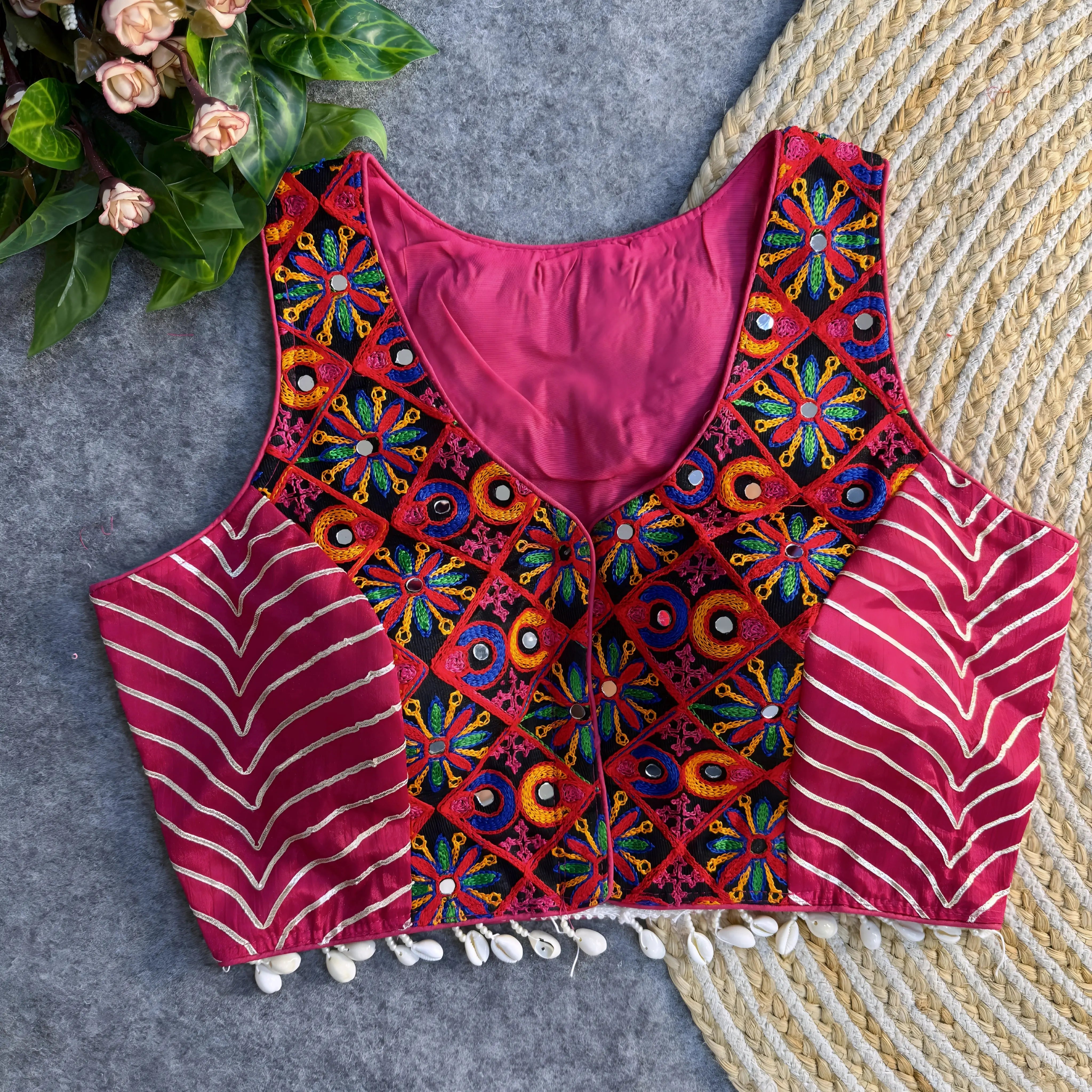 Sleeveless fuchsia ethnic blouse with vibrant mirror-work embroidery on the front, metallic chevron side panels, and cowrie-shell tassels along the hem, displayed on a grey backdrop with a woven mat and floral greenery