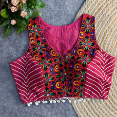 Sleeveless fuchsia ethnic blouse with vibrant mirror-work embroidery on the front, metallic chevron side panels, and cowrie-shell tassels along the hem, displayed on a grey backdrop with a woven mat and floral greenery