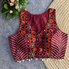 Sleeveless maroon ethnic blouse featuring colorful mirror work embroidery and metallic chevron side panels, displayed on a grey surface with a woven mat and foliage accents
