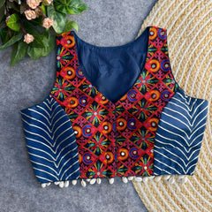 Sleeveless navy ethnic blouse with vibrant multicolored mirror-work embroidery on the front, metallic chevron side panels, and cowrie-shell tassels along the hem, displayed on a grey backdrop beside a woven mat and floral greenery