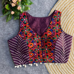 Sleeveless purple ethnic blouse accented with vibrant multicolored mirror-work embroidery on the front, metallic chevron side panels, and cowrie-shell tassels along the hem, displayed on a grey background next to a woven mat and floral greenery