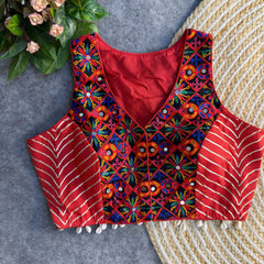 Sleeveless red ethnic blouse featuring multicolor mirror-work embroidery on the front panel, metallic chevron side panels, and cowrie-shell tassels along the hem, displayed on a grey surface beside a woven mat and floral greenery