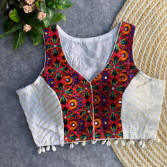 Sleeveless white ethnic blouse with vibrant mirror-work embroidery on the front, metallic gold chevron side panels, and cowrie-shell tassels along the hem, displayed on a grey backdrop beside a woven mat and greenery