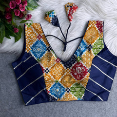 Traditional blouse featuring vibrant multicolor block print patterns with floral and geometric motifs in yellow, red, blue, and green, contrasted by navy blue silk side panels with golden stripes, and a deep V-neckline with patterned tasseled tie-back detail, displayed flat on a white fur background next to artificial pink flowers.