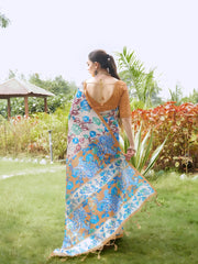 A woman models a graceful Beige Soft Tussar Silk Saree featuring a large, artistic, all-over Kalamkari print of blue and green floral motifs. She wears a rust-colored blouse and a chunky multi-colored beaded necklace, posing in a garden.