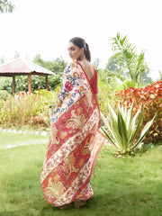 A woman models an elegant Beige Soft Tussar Silk Saree with a dense all-over print of multi-colored Kalamkari florals and vines. The saree has a wide, contrasting Deep Red border and pallu with traditional paisley print. She wears a matching red blouse and a chunky beaded necklace, posing outdoors in a bright garden setting.