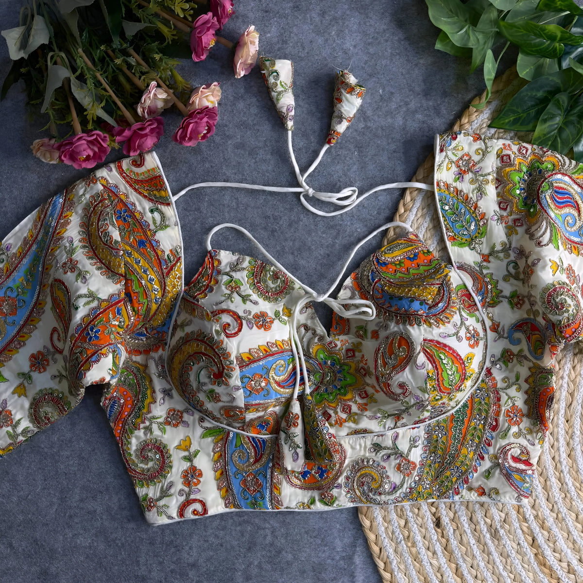 Multicolor paisley embroidered padded blouse with tie-up detail, displayed on a grey surface with artificial pink flowers and green leaves in the background.