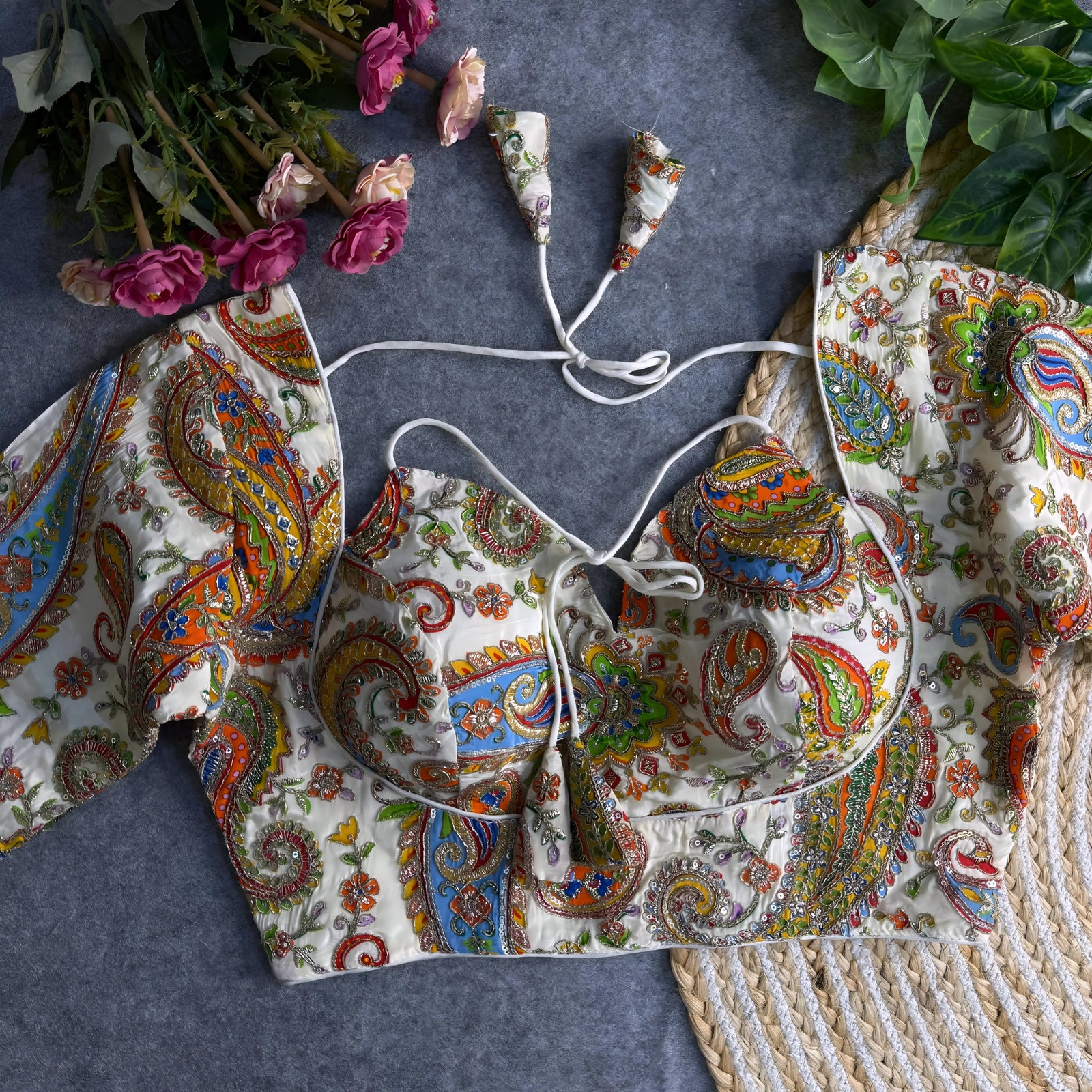 Multicolor paisley embroidered padded blouse with tie-up detail, displayed on a grey surface with artificial pink flowers and green leaves in the background.