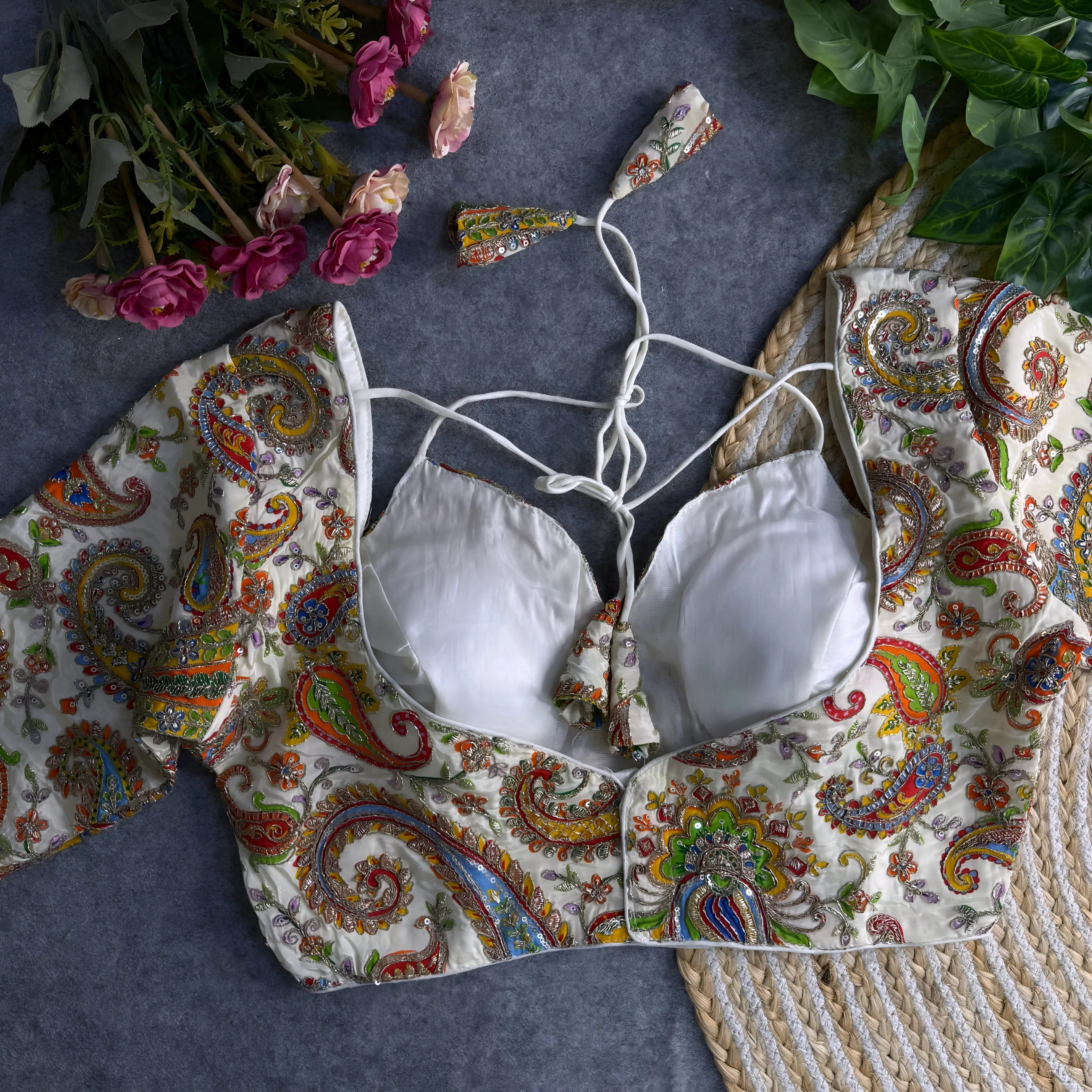 Multicolor paisley embroidered padded blouse with tie-up detail, displayed on a grey surface with artificial pink flowers and green leaves in the background