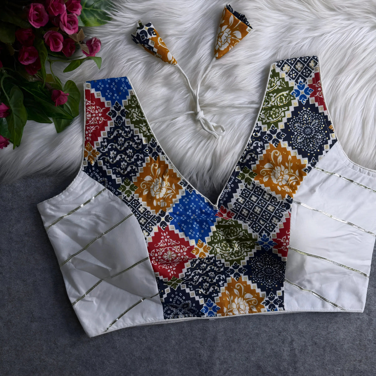 Traditional blouse featuring a multicolor patchwork pattern with intricate floral and geometric motifs, deep V-neckline, white silk side panels with gold accents, and patterned tasseled tie-back detail, displayed flat on a textured grey surface with artificial pink flowers and a white fur background.