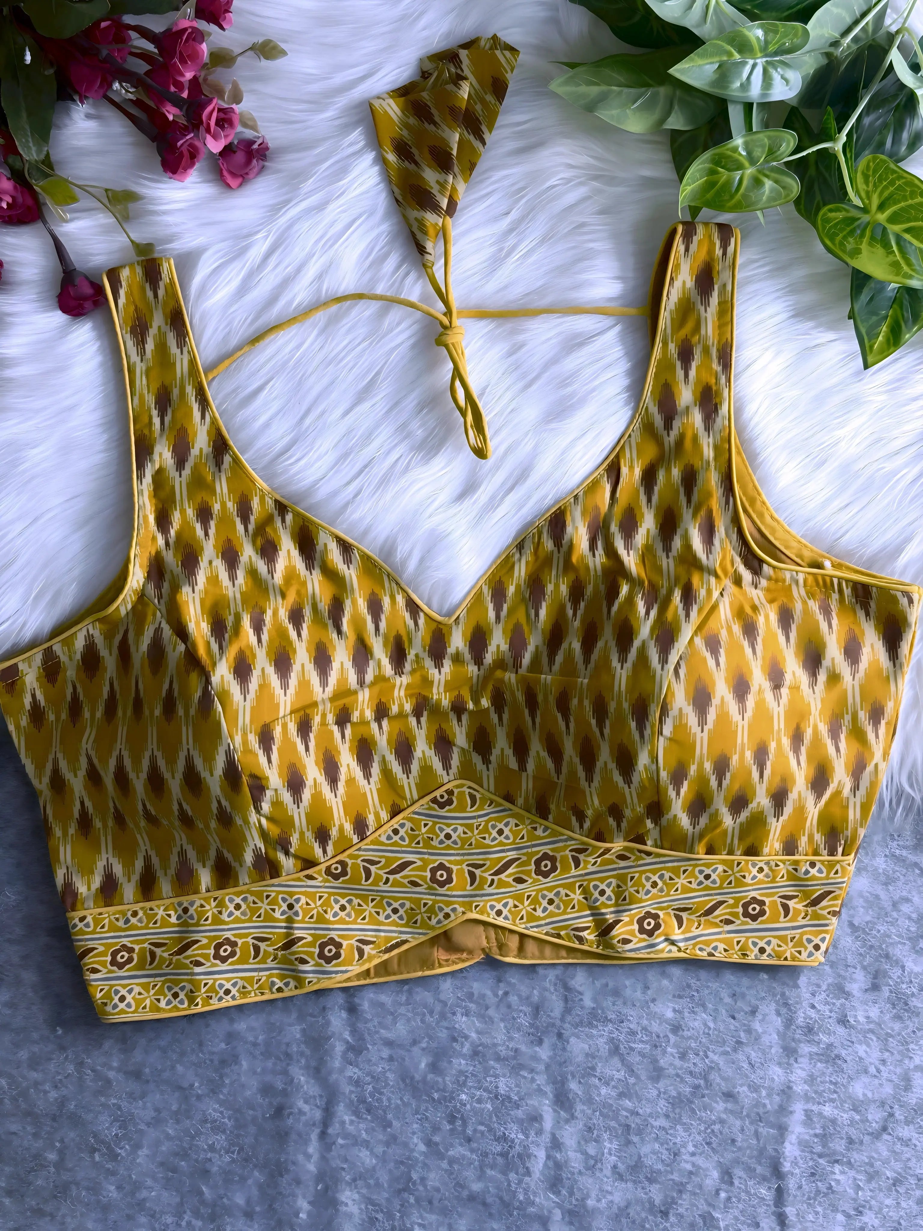 Mustard yellow silk blouse featuring traditional Ikat-inspired geometric print in brown and cream, wide shoulder straps, structured cups, and a decorative floral patterned border at the hem. Includes a matching tie-back detail with a petal-shaped tassel, displayed flat on a white fur background with pink artificial flowers and green foliage at the corners.