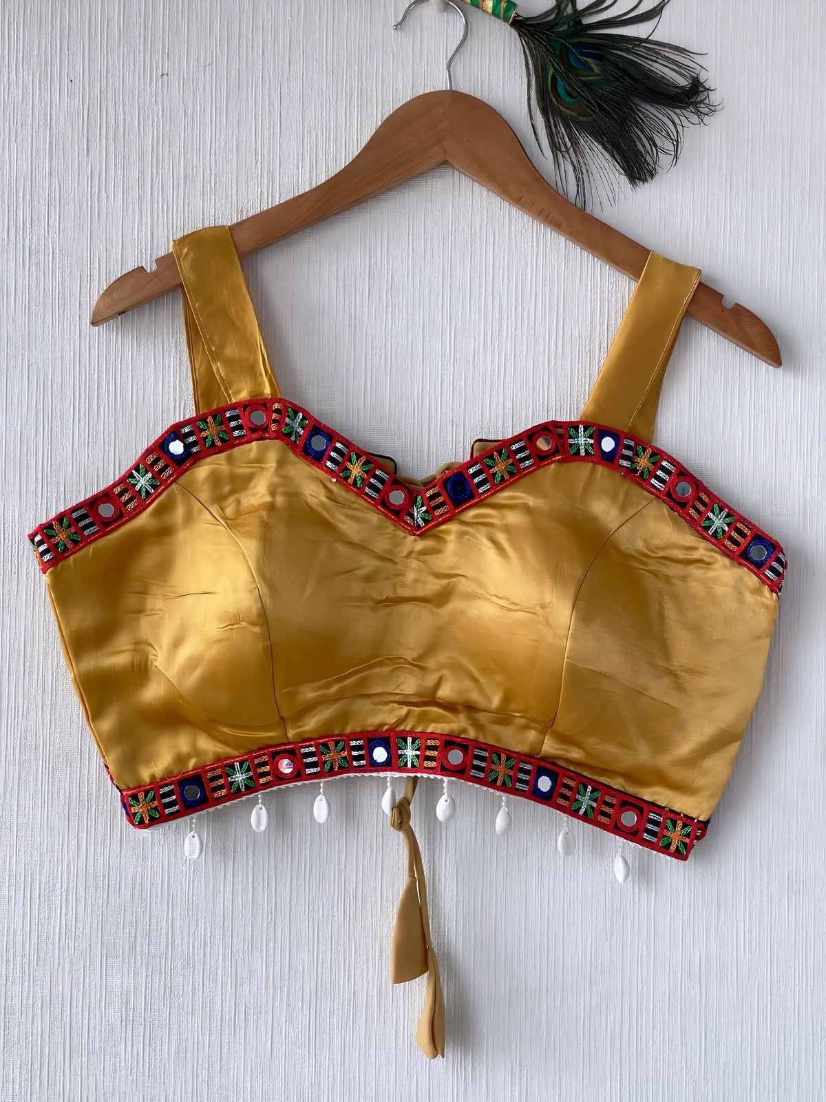 Mustard yellow ethnic blouse featuring colorful mirror work and embroidered geometric border in red, blue, and green, adorned with pearl drop tassels and a tie-up at the back, displayed on a wooden hanger against a textured off-white wall, creating a festive and traditional look.
