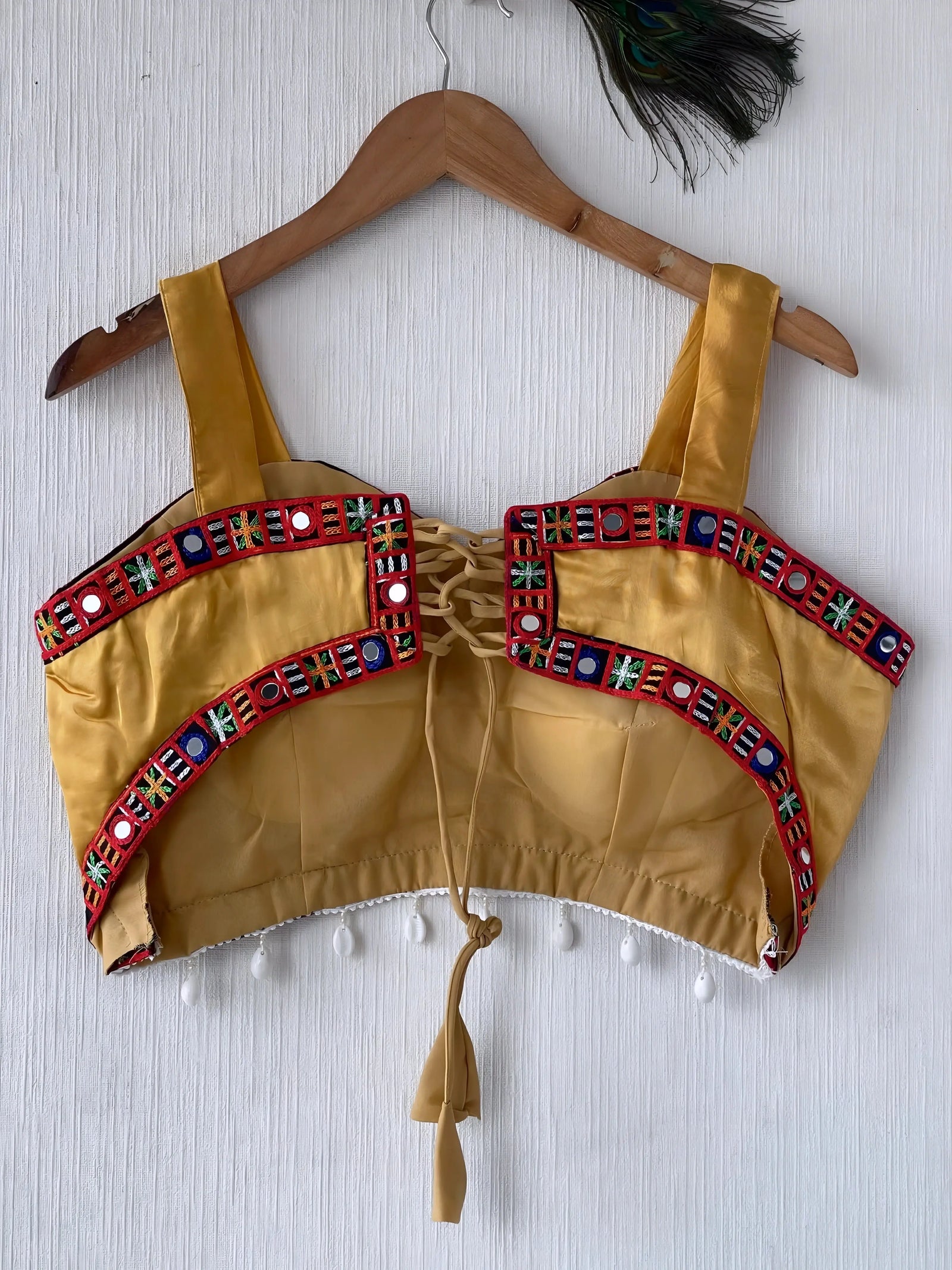 Mustard yellow ethnic blouse featuring colorful mirror work and embroidered geometric border in red, blue, and green, adorned with pearl drop tassels and a tie-up at the back, displayed on a wooden hanger against a textured off-white wall, creating a festive and traditional look.