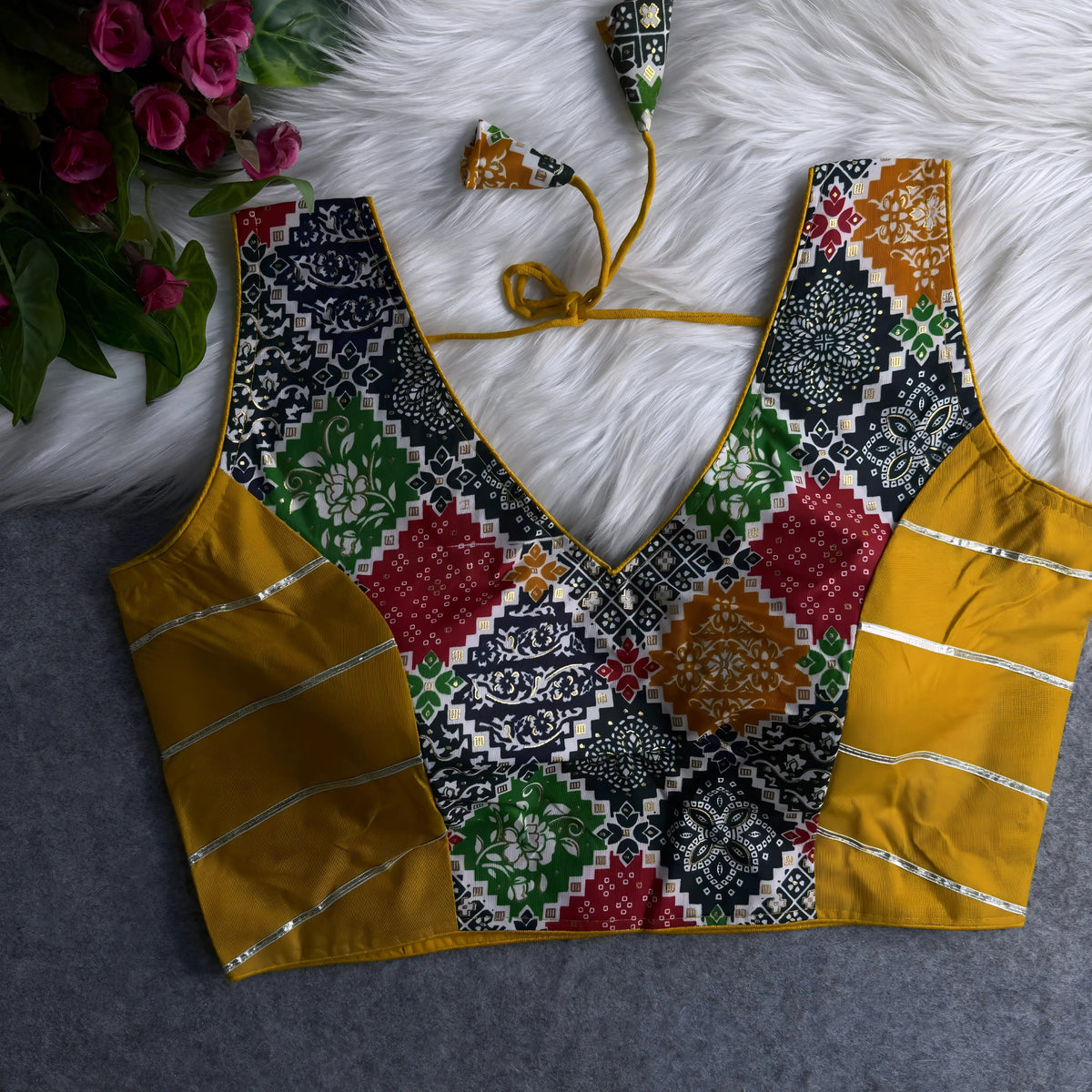 Mustard yellow and multicolor patchwork blouse with deep V-neckline, featuring intricate floral and geometric motifs, gold-accented side panels, and tasseled tie-back detail, displayed flat on a white fur surface with pink flowers in the corner.