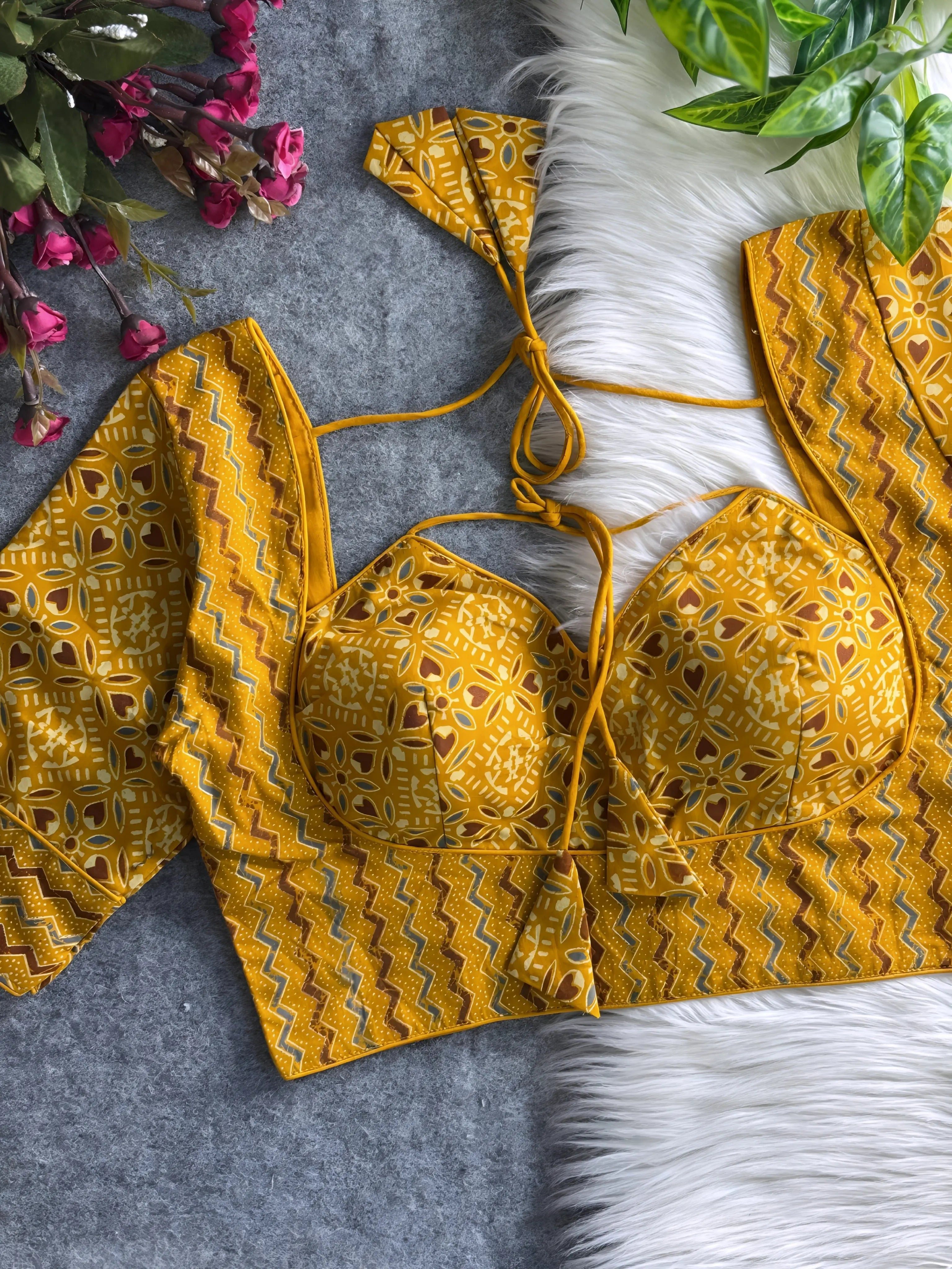 Mustard yellow printed padded blouse with geometric and floral patterns, displayed on a white fur surface with artificial flowers and green leaves in the background.