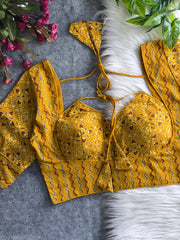 Mustard yellow printed padded blouse with geometric and floral patterns, displayed on a white fur surface with artificial flowers and green leaves in the background.
