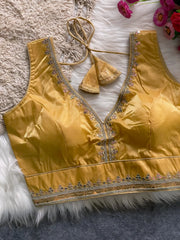 Traditional mustard yellow silk blouse featuring intricate gold sequin and thread embroidery along the neckline and hem, with a decorative tassel tie at the back, displayed flat on a soft fur and textured mat background with artificial pink flowers in the corner.