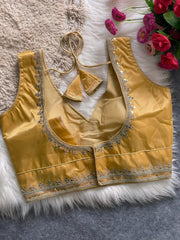 Traditional mustard yellow silk blouse featuring intricate gold sequin and thread embroidery along the neckline and hem, with a decorative tassel tie at the back, displayed flat on a soft fur and textured mat background with artificial pink flowers in the corner.