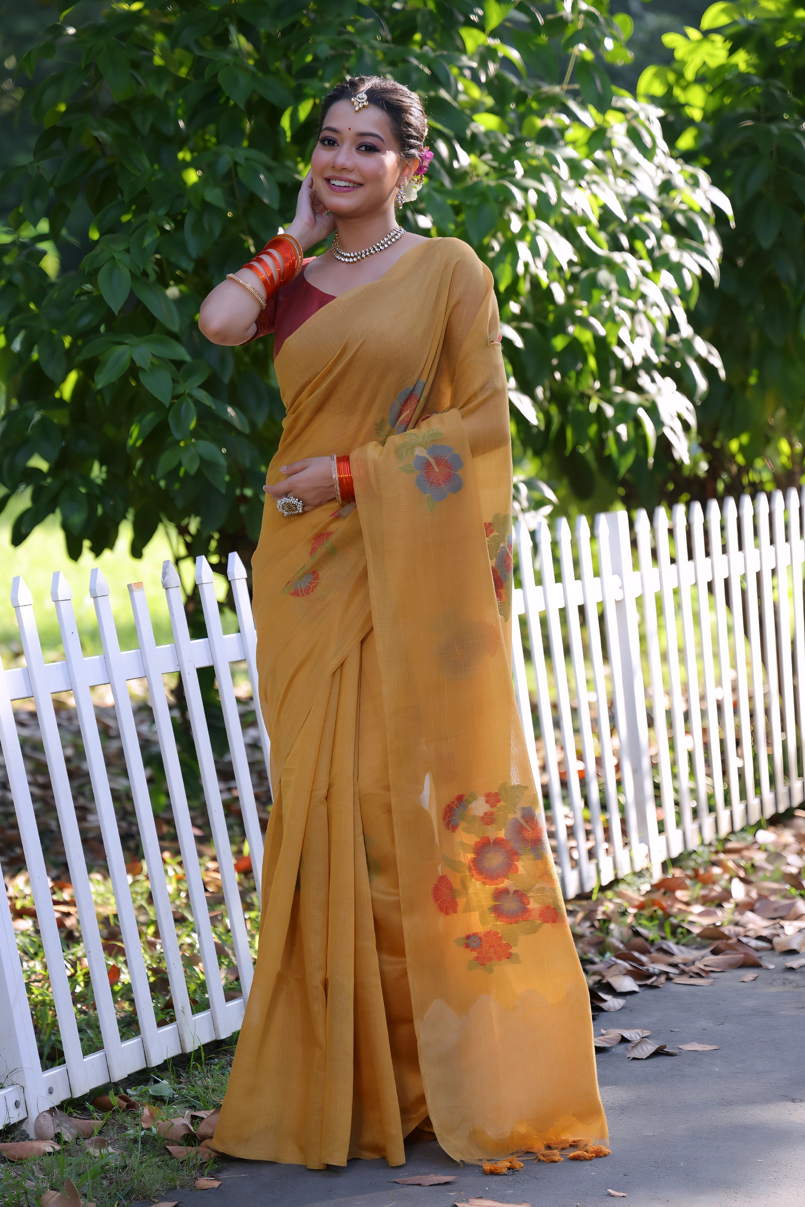 A woman models a vibrant Mustard Yellow Soft Muga Cotton Saree. The pallu features delicate colorful Resham floral weaving and tassels. She wears a maroon blouse and is accessorized with a white choker necklace and striking orange bangles, posing outdoors by a white fence.