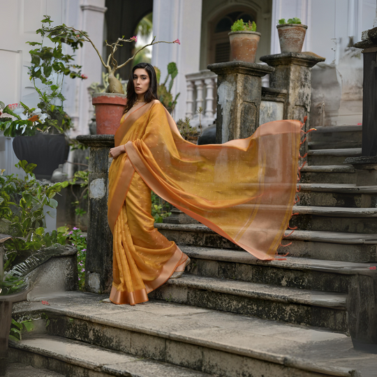 A woman models a stunning Golden Yellow Soft Silk Saree. The saree features subtle metallic motifs on the body and a wide, contrasting Copper/Rust Zari woven border. The lightweight fabric flows elegantly as she poses on a weathered stone staircase, wearing a delicate gold choker necklace.