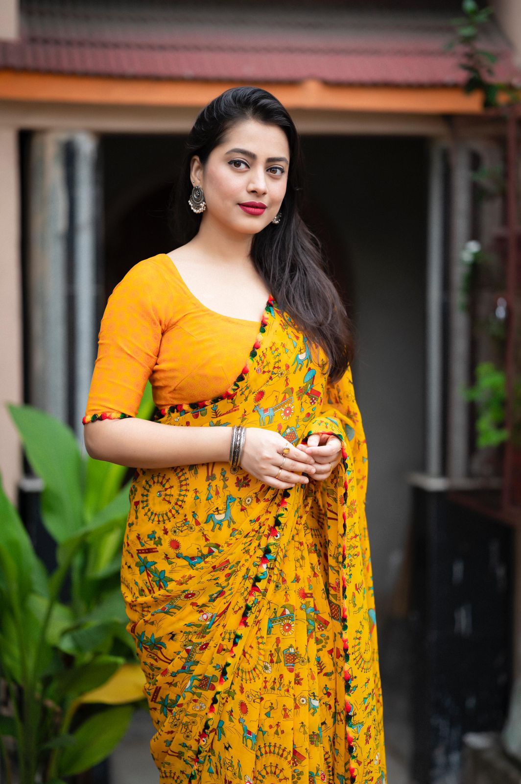 Woman in a mustard yellow Warli print saree with pom-pom border standing on a tiled patio with lush greenery and a traditional house in the background