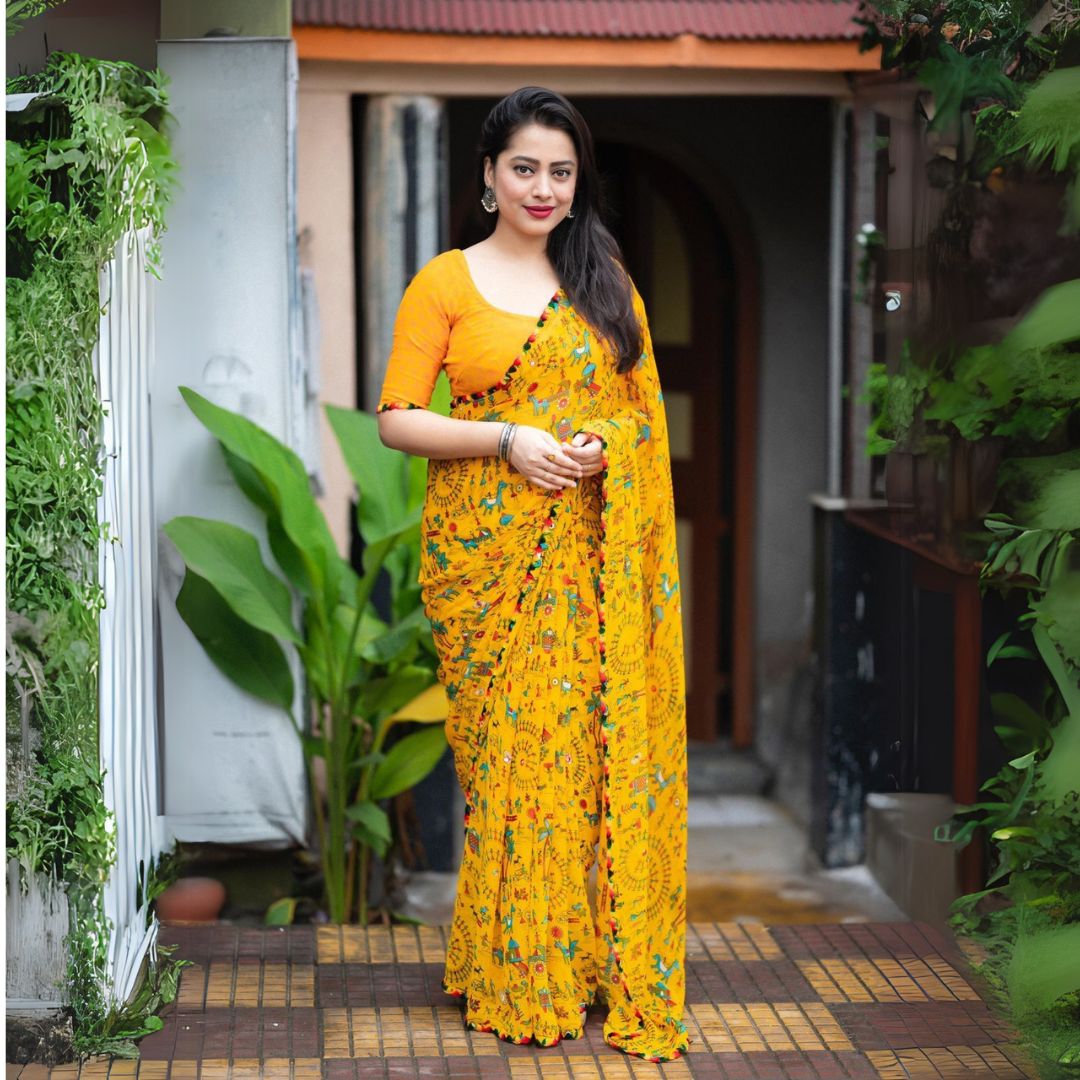 Woman in a mustard yellow Warli print saree with pom-pom border standing on a tiled patio surrounded by lush greenery and a traditional house backdrop