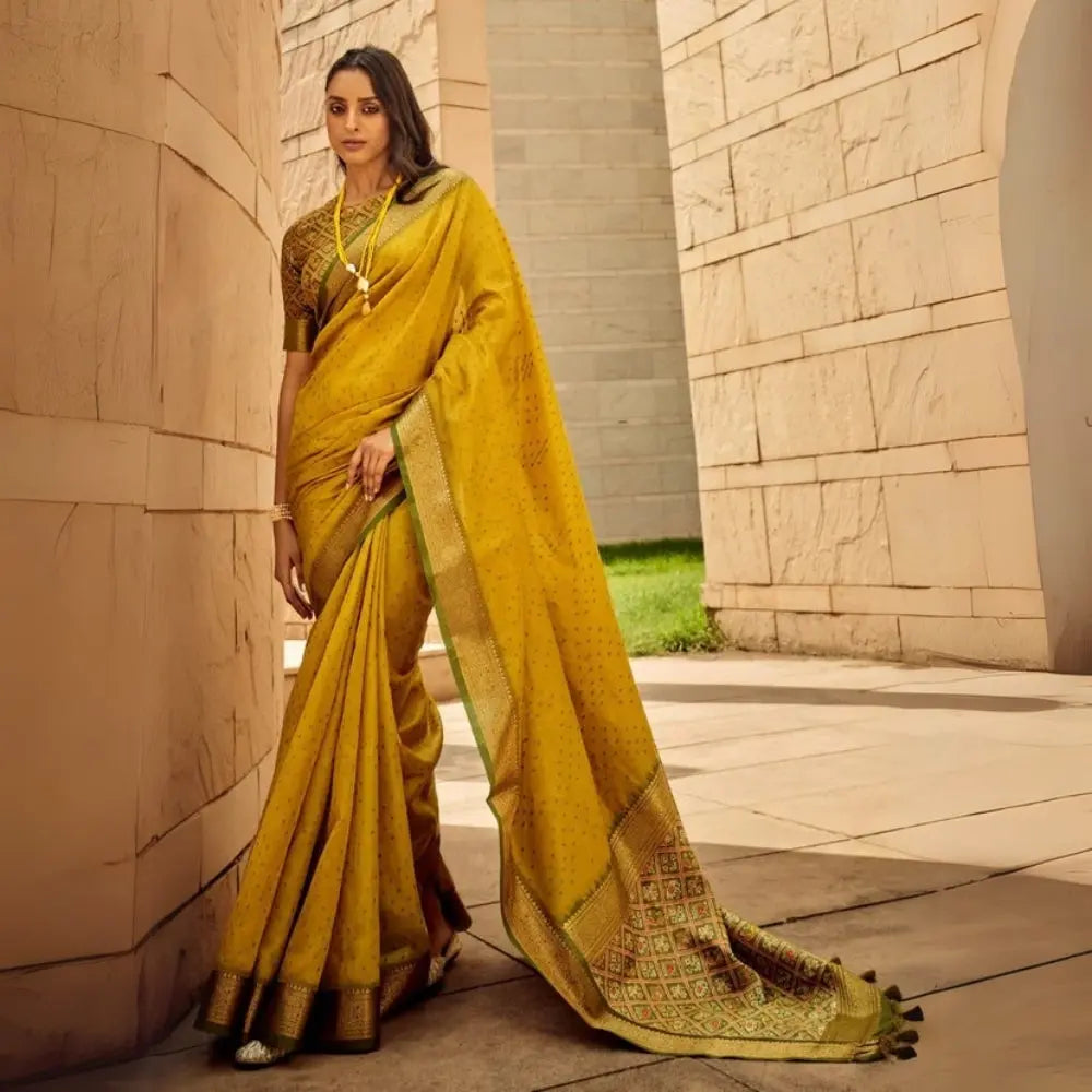 Woman wearing a mustard yellow saree with intricate zari work and green embroidered blouse, exuding traditional Indian elegance in a modern architectural setting
