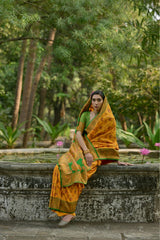 A person wearing a mustard yellow saree with intricate zari work, standing in front of a stone structure with a lush green background and pink flowers.