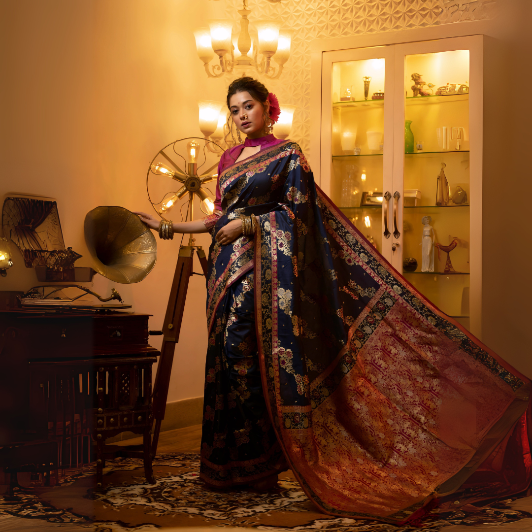 A woman models a luxurious Navy Blue Soft Banarasi Silk Saree. The saree features dense multi-colored Zari weaving of florals across the body and a wide pallu with heavy gold and deep red/maroon weaving. She wears a high-neck Fuchsia Pink blouse and gold jewelry, posing in a dramatic, opulently lit vintage setting.