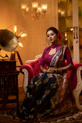A woman models a luxurious Navy Blue Soft Banarasi Silk Saree. The saree features dense multi-colored Zari weaving of florals across the body and a wide pallu with heavy gold and deep red/maroon weaving. She wears a high-neck Fuchsia Pink blouse and gold jewelry, posing in a dramatic, opulently lit vintage setting.