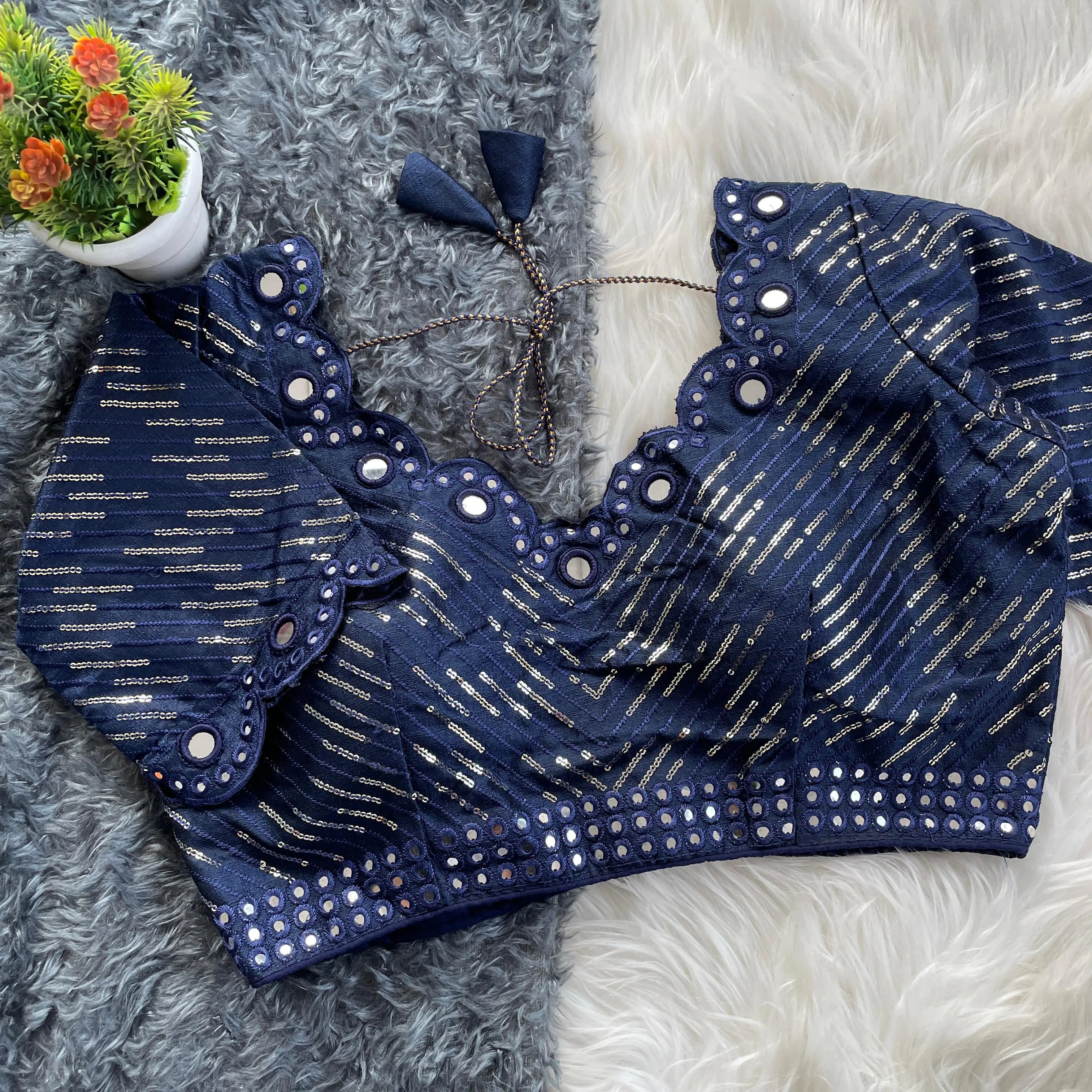 Navy blue blouse with silver sequin stripes, scalloped neckline embellished with mirror work, decorative sleeve borders, and tie-up tassels, displayed on a textured grey and white fur background with a small potted plant nearby.